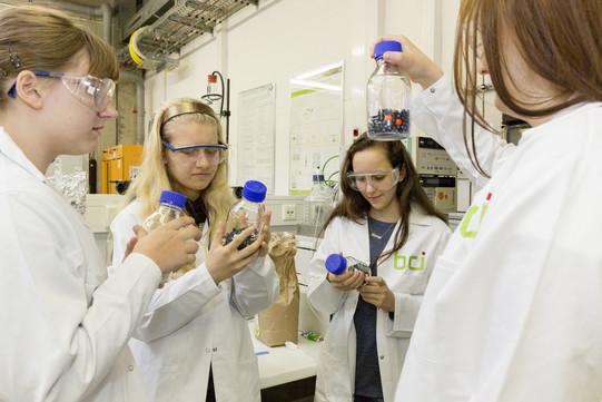 Vier Mädchen sind in einem Labor, tragen weiße Kittel und halten Proben in den Händen