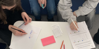 Von oben fotografiert: Zwei Personen sitzen an einem weißen Tisch und schreiben auf Papier mit Stiften, auf dem Tisch liegen bunte Notizzettel und ausgedruckte Blätter mit Diagrammen