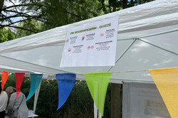 Bunte Wimpelkette vor einem weißen Pavillon mit einem Plakat zur "Die Besserwisserin - Quizze"-Aktion im Freien unter Bäumen.