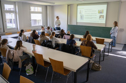 Schülerinnen sitzen in einem hellen Seminarraum an Tischen und hören zwei Vortragenden zu, die vor einer Tafel mit projizierter Präsentation stehen