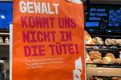 An orange paper bag with white and red text stands in a bakery in front of a shelf with baked goods. The bag reads in large letters: 'VIOLENCE DOES NOT GO INTO OUR BAG!'. Below is a white silhouette profile of a woman's face. Further down, it says: 'An action for the International Day for the Elimination of Violence against Women on November 25'. Contact details for a women's counseling center in Dortmund, including phone number and website, are printed. The bag is part of a campaign against violence toward women and girls. Various types of bread and pastries are visible in the background.
