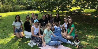 Die Mentees und Mentorinnen (insgesamt 14 Personen) sitzen auf einer Wiese unter einem Baum und lächeln in die Kamera. 