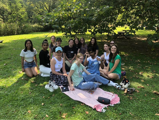 The mentees and mentors (14 people in total) are sitting in a meadow under a tree and smiling at the camera. 