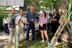 Gruppe von Menschen steht und unterhält sich im Schatten eines Baums bei einem Sommerfest mit einem Tisch im Hintergrund und einem Staffelei-ähnlichen Objekt im Vordergrund.