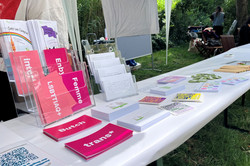 Langer Tisch unter einem weißen Pavillon mit Informationsmaterialien zu LGBTQ+-Themen und zwei Personen im Hintergrund in einem Garten