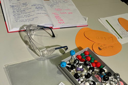 Tisch mit geöffnetem Notizbuch, Schutzbrille, Metallbox mit bunten Reißzwecken, orangefarbenen Papierstücken und mehreren Zetteln mit handschriftlichen Notizen.