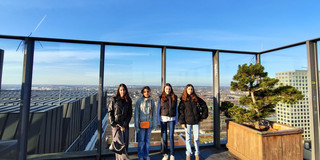 Four girls stand next to each other on the viewing platform of the Dortmunder U and look into the camera. Behind the large glass pane, a wide view stretches out over Dortmund. The sky is bright blue and almost cloudless.