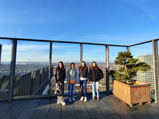 Auf der Aussichtsplattform des Dortmunder U#S stehen vier Mädchen nebeneinander und schauen in die Kamera. Hinter der großen Glasscheibe erstreckt sich ein weiter Blick über Dortmund. Der Himmel ist strahlend Blau und nahezu wolkenlos.