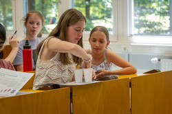Zwei Mädchen sitzen im Hörsaal. Aus weißen Plastikbechern verkosten sie verschiedene Schokoladensorten.