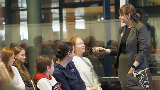 Woman in a gray blazer and green skirt holds a microphone in front of a seated group of young people in an interior room with glass walls.