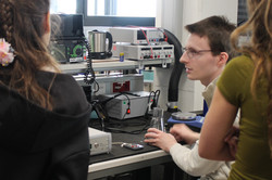 Mehrere Mädchen stehen in einem Technikraum der TU Dortmund und beobachten eine Person, die an einem Computerarbeitsplatz mit technischen Geräten sitzt.
