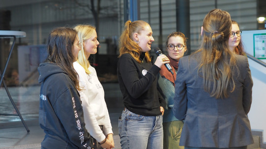 Gruppe von fünf weiblichen Personen stehen in einem Innenraum, eine Frau hält ein Mikrofon, im Hintergrund Glasfassade und Monitor.