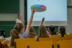 Von hinten fotografiert: Mädchen sitzen in einem Hörsaal, zwei strecken eine Hand in die Höhe. Vor ihnen steht die Workshopleiterin vor einer Leinwand mit einer Präsentation. 