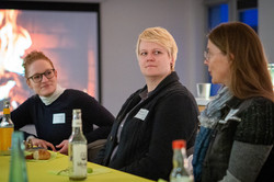 Drei Personen sitzen an einem Tisch mit mehreren Flaschen und einem Brot, im Hintergrund ein Bildschirm mit einem Feuerbild.