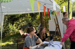 Zwei Frauen sitzen an einem mit Laptop und Beamer ausgestatteten Tisch unter einem weißen Pavillon mit bunten Wimpeln, ein Mann spricht sie an, im Hintergrund Bäume und weitere Personen.