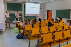 Zwölf Mädchen sitzen in einem Hörsaal. Vor einer Leinwand stehen zwei Personen und halten einen Vortrag. Auf der Leinwand steht "Irreführende Grafiken".