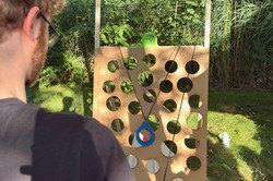Person mit schwarzem T-Shirt spielt im Garten ein Geschicklichkeitsspiel mit Seilen und einem Brett mit runden Löchern.