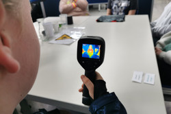 A girl holds a thermal camera in her hand and points it at another girl. The outline of the body appears on the screen in orange tones.