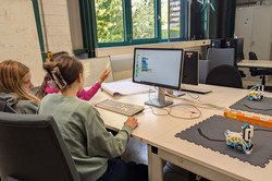 Drei Mädchen sitzen vor einem Computer. Neben ihnen auf dem Tisch steht ein Logo-Roboter auf einem Testfeld. 