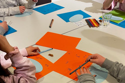 Kinder sitzen an einem Tisch und zeichnen auf bunten Papierbögen mit Stiften.