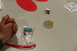 Gefaltete Hände auf einem Tisch neben einem durchsichtigen Plastikbeutel mit kleinen Gegenständen, einem kleinen Würfel, einem runden Behälter und roten Papierkreisen mit handgeschriebenem Text