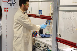 A man dressed in a white protective coat, safety goggles and blue rubber gloves stands in front of a window in a laboratory. Behind it are glasses and equipment.