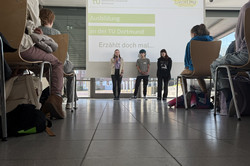 ‘Tell me about it’ is written on a screen.Three trainees stand in front of it. The Girls Day participants are sitting on chairs. 