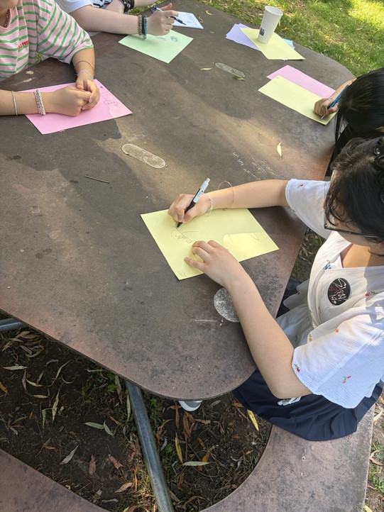 Mehrere Personen sitzen an einem runden Außentisch und schreiben auf verschiedenfarbige Blätter Papier.