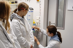 A schoolgirl fills a liquid from a large container into a small tube. Two other girls are standing next to her. All three are wearing white coats and protective goggles. 