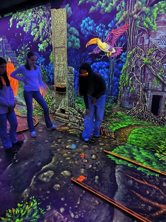 Ein Mädchen spielt Minigolf auf einer Schwarzlicht-Minigolf Bahn, zwei weitere sehen zu.
