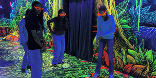 Ein Mädchen spielt auf einer Schwarzlicht-Minigolf Bahn, die mit Dschungelpflanzen in Neonfarben dekoriert ist. Drei weitere Mädchen sehen zu.