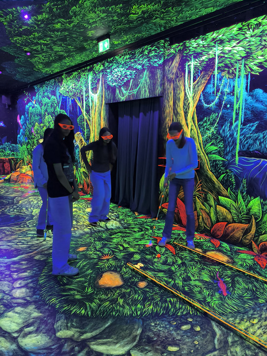 Ein Mädchen spielt auf einer Schwarzlicht-Minigolf Bahn, die mit Dschungelpflanzen in Neonfarben dekoriert ist. Drei weitere Mädchen sehen zu.