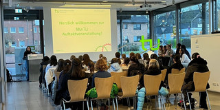 Several people sit on wooden chairs in a bright room with large windows listening to a speaker at a podium in front of a screen displaying the project welcome message 'Herzlich willkommen zur MinTU Auftaktveranstaltung'.