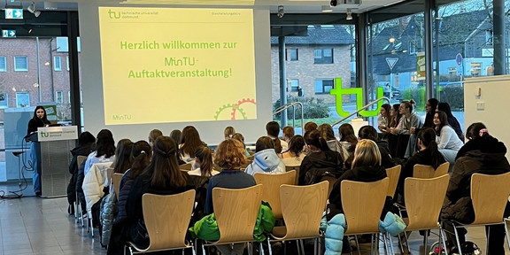 Mehrere Personen sitzen in einem hellen Raum mit großen Fenstern auf Holzstühlen und hören einer Rednerin am Rednerpult zu, die vor einer Leinwand mit der Projekteinladung 'Herzlich willkommen zur MinTU Auftaktveranstaltung' steht.
