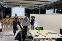 Innenraum mit mehreren Personen, die in kleinen Gruppen stehen und sitzen, Tische mit Papieren und Stiften, im Hintergrund eine Leinwand mit Projektion