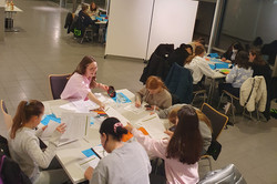 Mehrere Gruppen von Personen sitzen an Tischen in einem großen, modernen Raum mit Glaswänden und arbeiten an Papieren und Laptops.