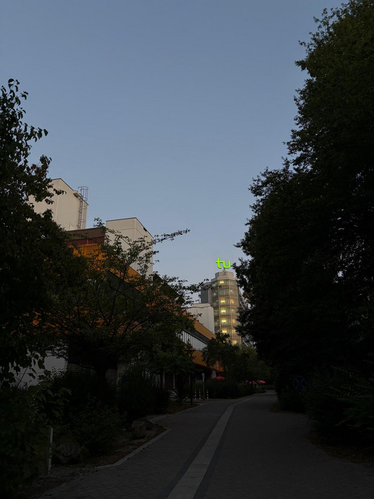 Ein schmaler Weg verläuft zwischen dicht bewachsenen Bäumen und Gebäuden. Im Hintergrund ragt ein mehrstöckiges Gebäude mit beleuchteten Fenstern in den Himmel. Auf dem Dach des Gebäudes leuchtet ein grünes Logo mit den Buchstaben 'tu'. Der Himmel ist klar und hellblau, es sind keine Menschen zu sehen.