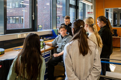 The workshop leader is sitting at a lathe. Three girls stand around him and listen to him. 