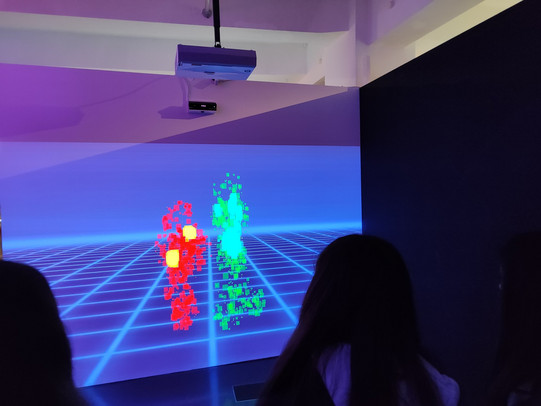 Auf einer Leinwand werden in zwei verschiedenen Farben Figuren aus Pixeln abgebildet. Davor stehen drei Mädchen und schauen auf die Leinwand.