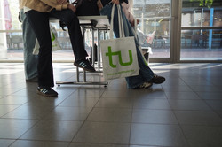 Mehrere Personen stehen in einem Innenraum auf gefliesten Boden, eine Person hält eine weiße Tasche mit grünem "tu"-Logo und der URL "www.tu-dortmund.de".