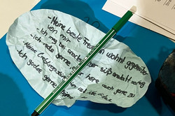 Blatt mit handschriftlichem Text auf blauem Untergrund, darauf liegt ein grüner Buntstift, daneben ein Smartphone mit farbigem Muster.