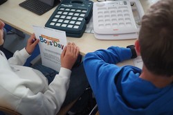 Zwei Jungen sitzen an einem Tisch mit technischen Geräten und halten jeweils ein Blatt mit der Überschrift 'Boys'Day' in der Hand.