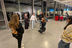 Five girls stand in a circle, a sixth girl sits in the centre and pantomimes something. 