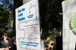 Plakat mit Erklärungstext zur "Leaky Pipeline" an einem grauen Pfosten im Freien, im Hintergrund Menschen und Bäume.