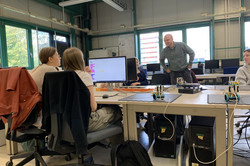 Zwei Mädchen sitzen vor einem Computer. Ein Mann steht ihnen gegenüber und lehnt sich in Richtun Schreibtisch.