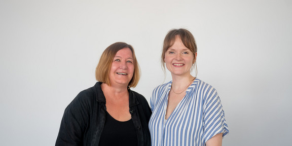 Two women stand next to each other in front of a white wall and smile into the camera.