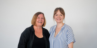 Two women stand next to each other in front of a white wall and smile into the camera.