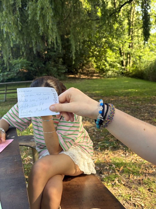 Eine Person sitzt auf einer Bank im Freien und hält sich mit einer Hand das Gesicht verdeckt, eine andere Hand hält eine kleine Karte mit handschriftlichem Text in den Vordergrund.