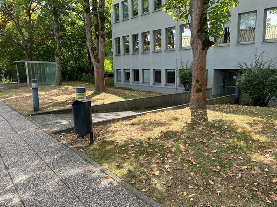 Foto von einem grauen Gebäude mit vielen Fenstern. Vor dem Gebäude verläuft ein Weg, von dort führt eine Rampe zu einer Tüt. Links und rechts von der Rampe befindet sich Wiese.nter zu einer Tür