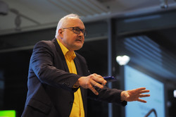 An older man in a dark jacket and yellow shirt gestures with both hands, holding an ultrasound device in his right hand, in front of a window in an event room.